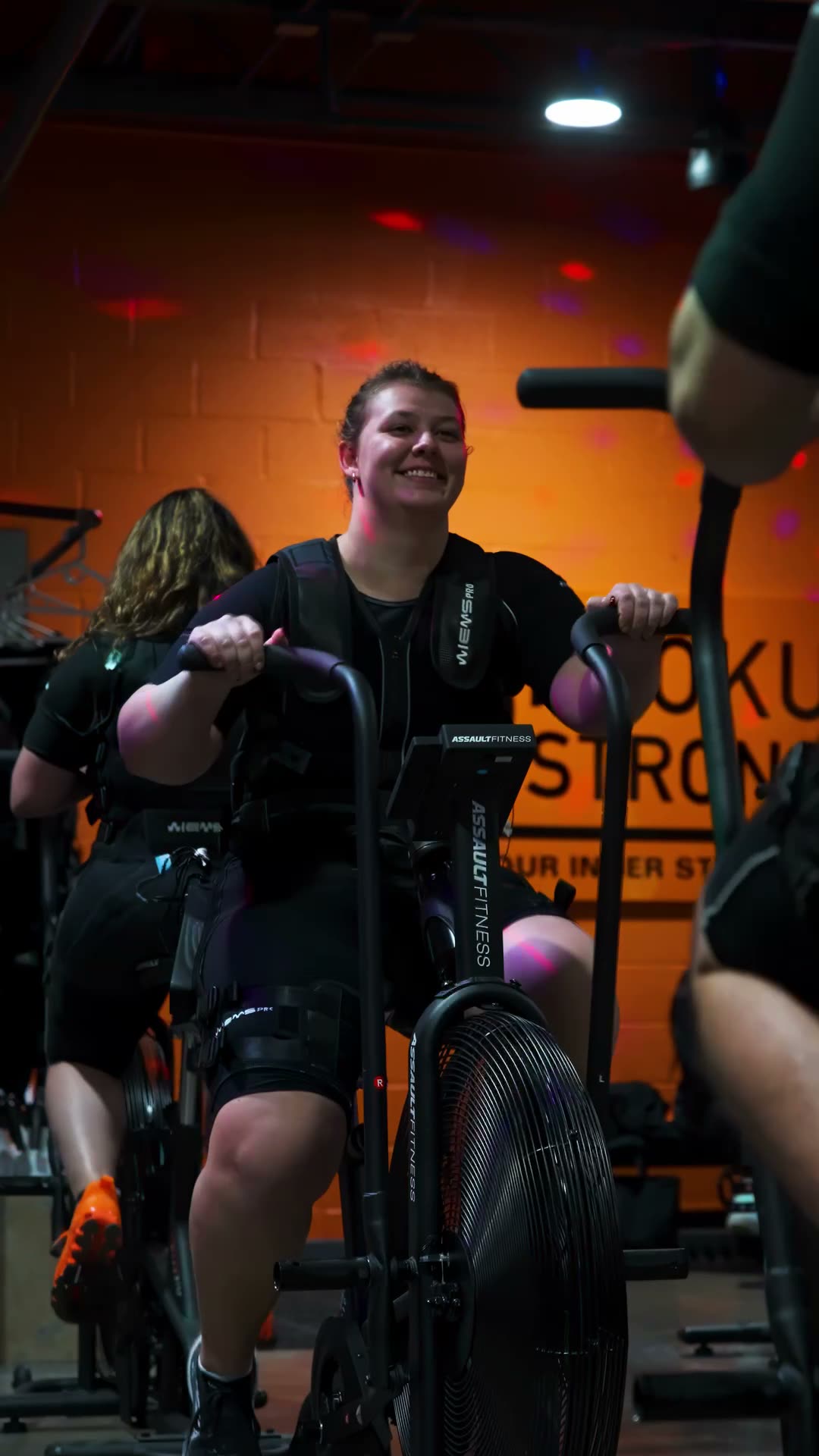A member in a Njoku Strong EMS training suit driving a studio air bike under club lighting, against the orange NJOKU STRONG wall in the Westlake studio.