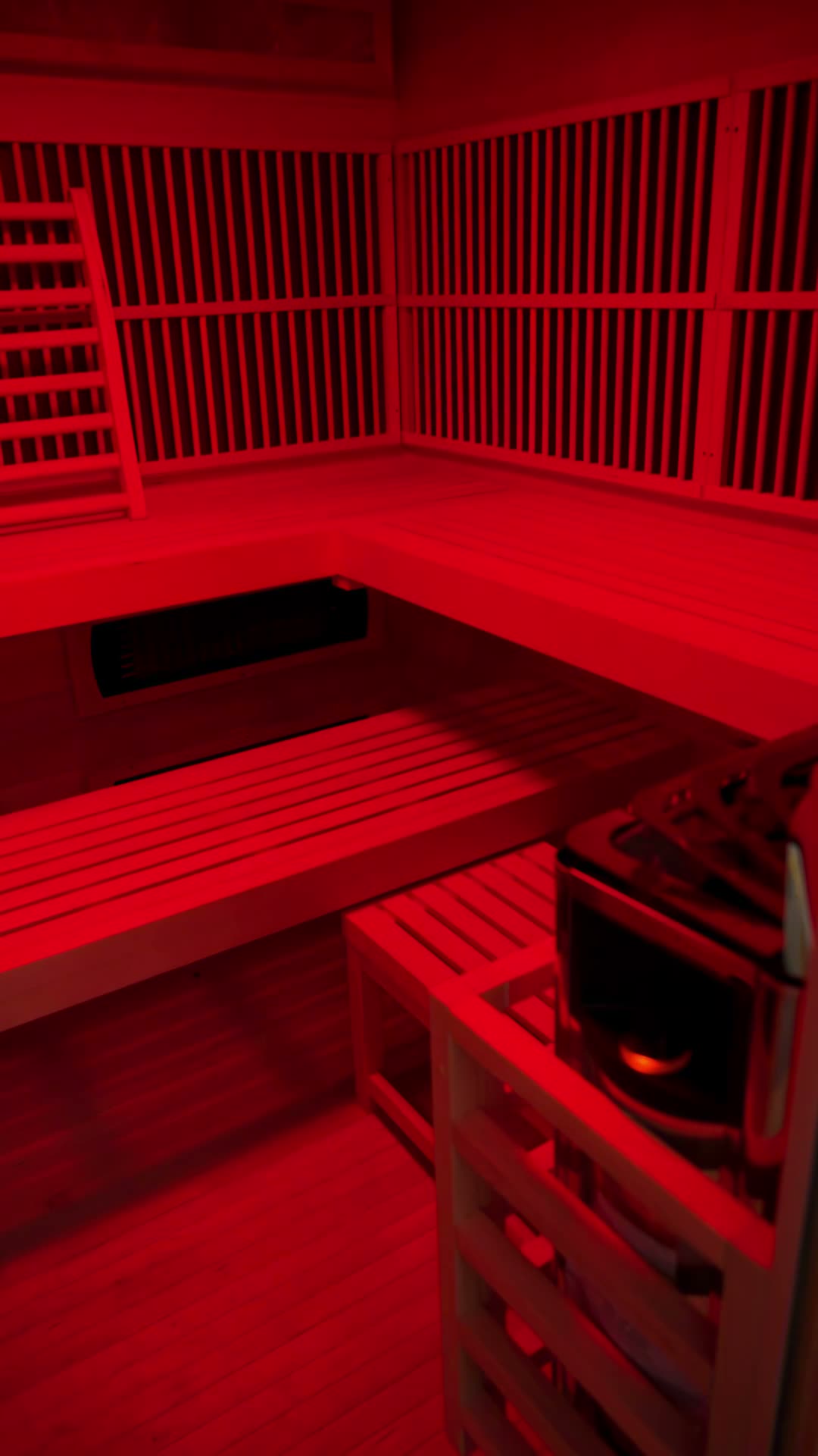Inside the infrared sauna cabin: an L-shaped cedar bench, slatted infrared heater panels along the walls and a towel caddy, the whole cabin lit deep red.