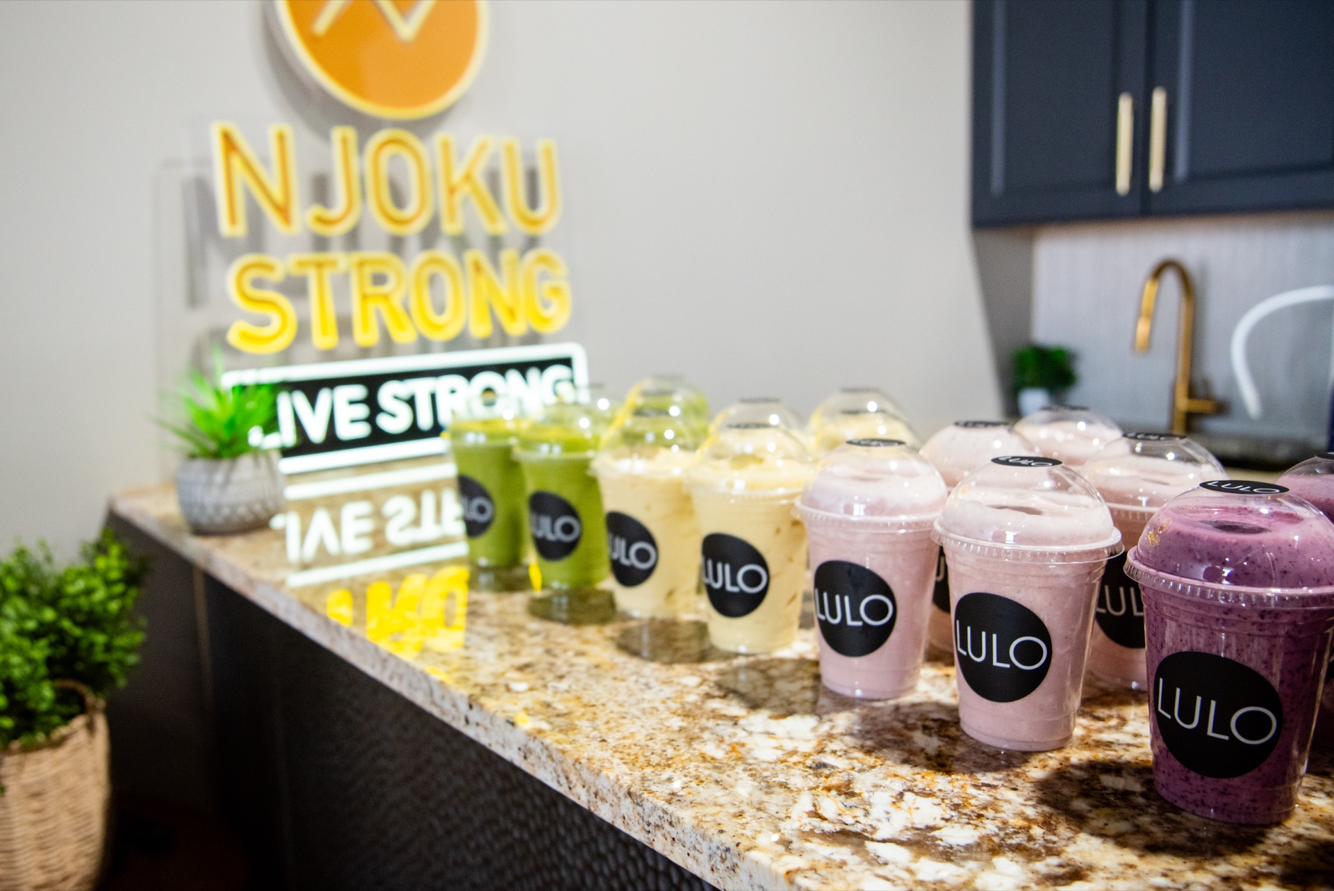 The smoothie bar counter at Njoku Strong: rows of smoothies — green, cream, strawberry, and blueberry — on a granite counter beneath the neon NJOKU STRONG, LIVE STRONG sign.